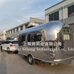 Stainless steel Airstream Allie pop-up caravan parked outside an industrial building, shiny reflective exterior and tow hitch