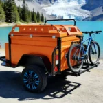 Orange Box Buddy mini camper trailer with rear bike rack parked by turquoise alpine lake, off-road compact caravan