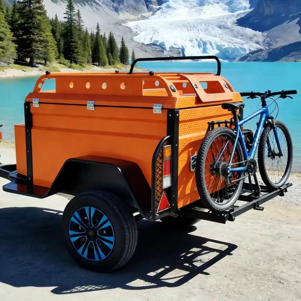 Orange Box Buddy mini camper trailer with rear bike rack parked by turquoise alpine lake, off-road compact caravan