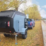 Cabin Clam (CODE 2679) off-road camper trailer hitched to a blue truck parked by a lake during an autumn afternoon.