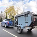 Cabin Clam (CODE 2679) pop-up off-road caravan being towed by a blue pickup truck on a scenic highway with a rainbow.