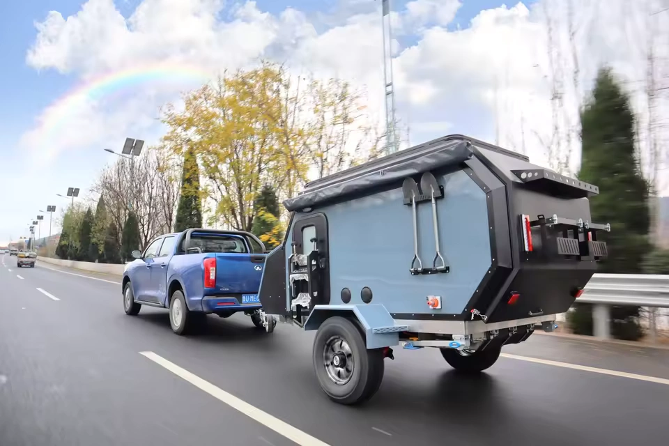 Cabin Clam (CODE 2679) pop-up off-road caravan being towed by a blue pickup truck on a scenic highway with a rainbow.
