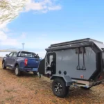 The Cabin Clam (CODE 2679) off-road camper trailer towed by a blue pickup truck near a scenic lakeside road view.
