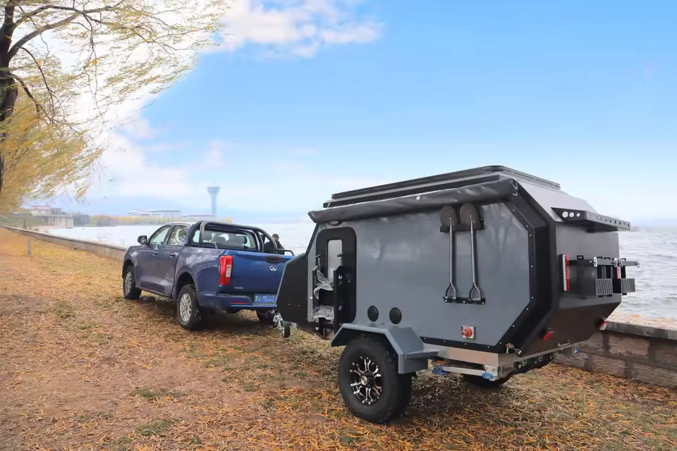 The Cabin Clam (CODE 2679) off-road camper trailer towed by a blue pickup truck near a scenic lakeside road view.