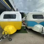 Two compact "Capsule" pop-up caravans in yellow and blue parked side-by-side at a caravan dealer lot, Molly Moulds model