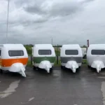 Row of four compact Molly Moulds mini caravans in orange, green, grey and white parked on wet asphalt at dealership