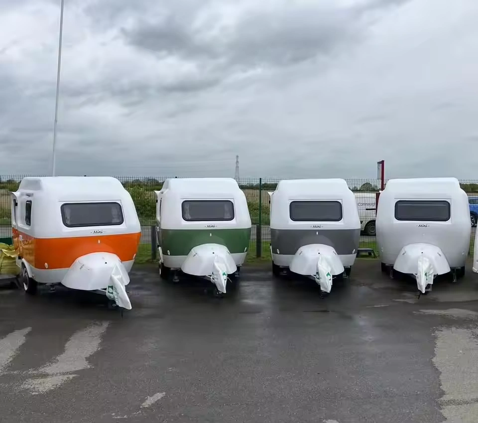Row of four compact Molly Moulds mini caravans in orange, green, grey and white parked on wet asphalt at dealership