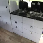 Roming Romeo camper trailer interior featuring white storage drawers, stainless sink, two-burner stove and wood-look flooring