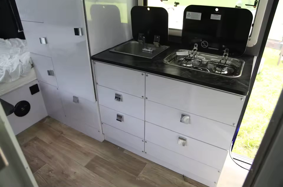 Roming Romeo camper trailer interior featuring white storage drawers, stainless sink, two-burner stove and wood-look flooring