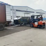 Rugged Roy off-road teardrop trailer in olive drab being forklift-loaded into a shipping container at factory yard