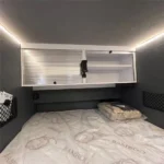 Modern interior of the Swiss Army Andy caravan featuring a comfortable queen-size bed and overhead storage cabinets.