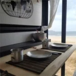 Van conversion interior dining setup with stainless steel plates, cups and bowl by window overlooking sandy beach view