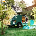 The Cody Compact mini caravan set up in a grassy campsite with an open door, rooftop tent, and side awning extended.