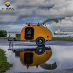 Yellow Cody Compact mini teardrop camper trailer with an open rear hatch parked near a coastal harbor scene.